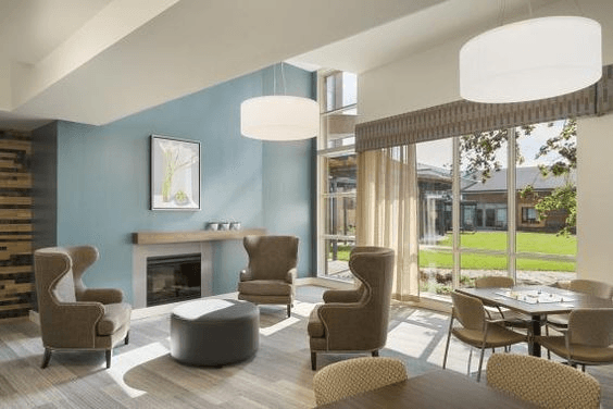A bright, comfortably furnished lounge in one of our residential care homes, with armchairs, a fireplace, and large windows